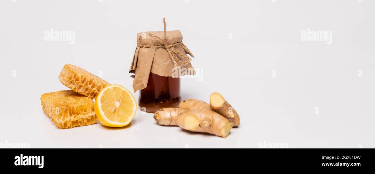 vaso di miele coperto di carta artigianale vicino al limone fresco, radice di zenzero e nido d'ape su bianco, striscione Foto Stock