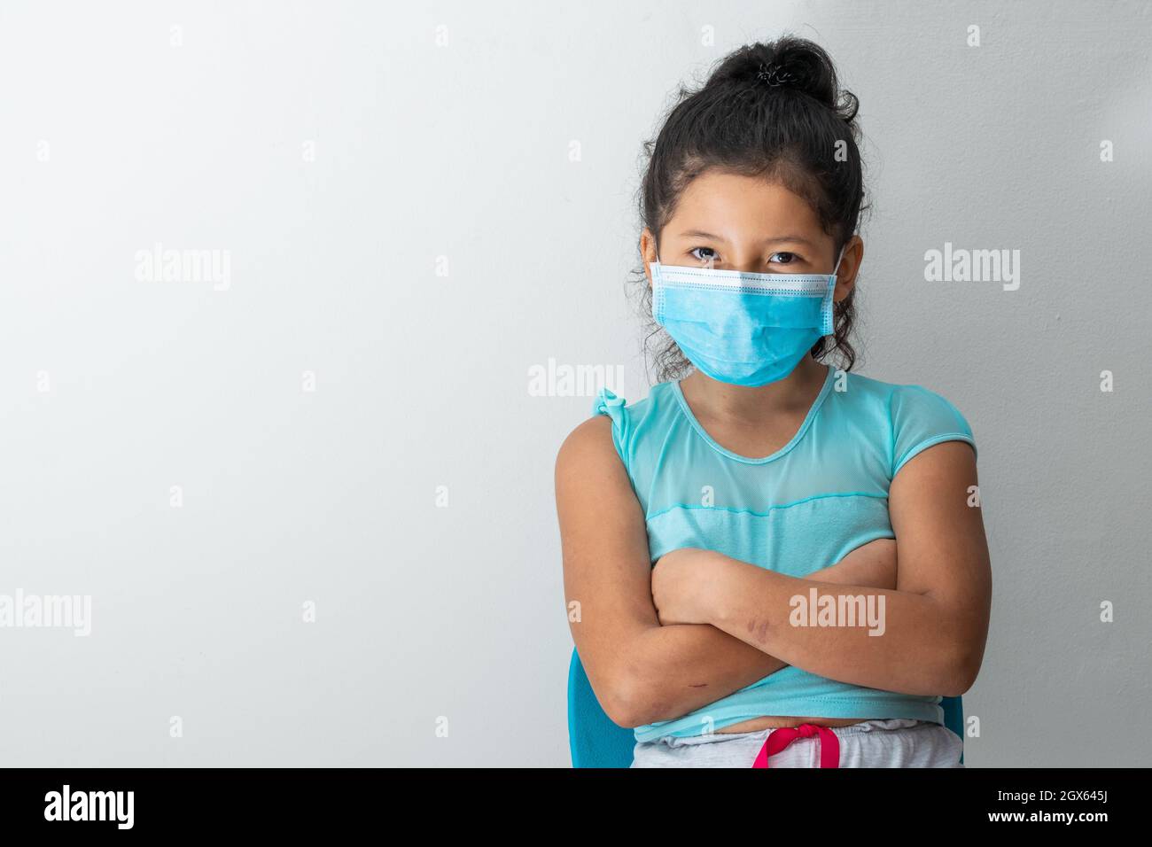 bambina (8 anni) che guarda la macchina fotografica con un volto maligno, seduta con braccia incrociate e maschera chirurgica blu. Medico, farmaceutico e s. Foto Stock
