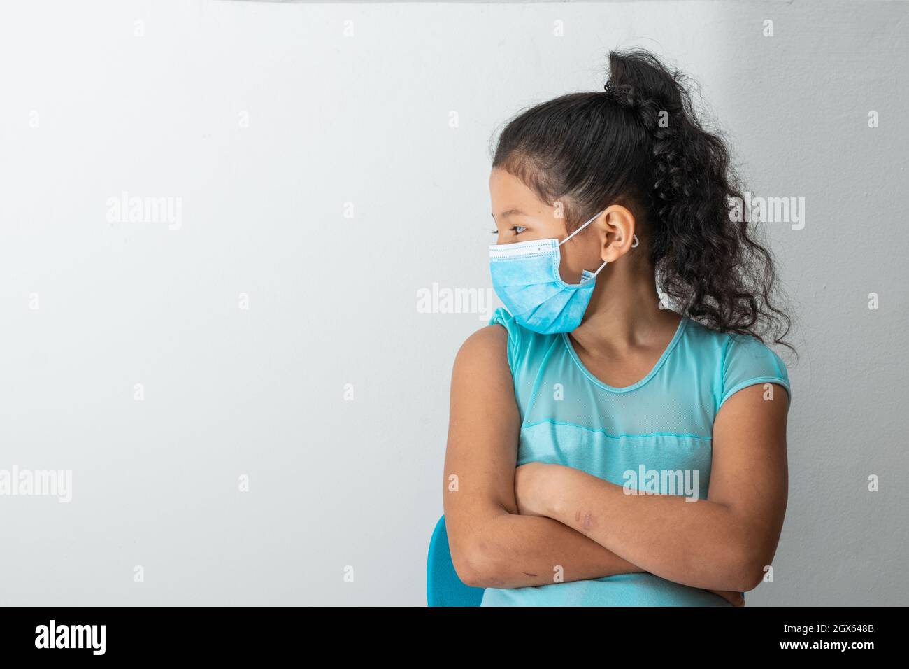 bambina (8 anni) che guarda a destra sorridendo, allegro sguardo, seduta con le braccia incrociate e maschera chirurgica blu. Medico, farmaceutico e s. Foto Stock