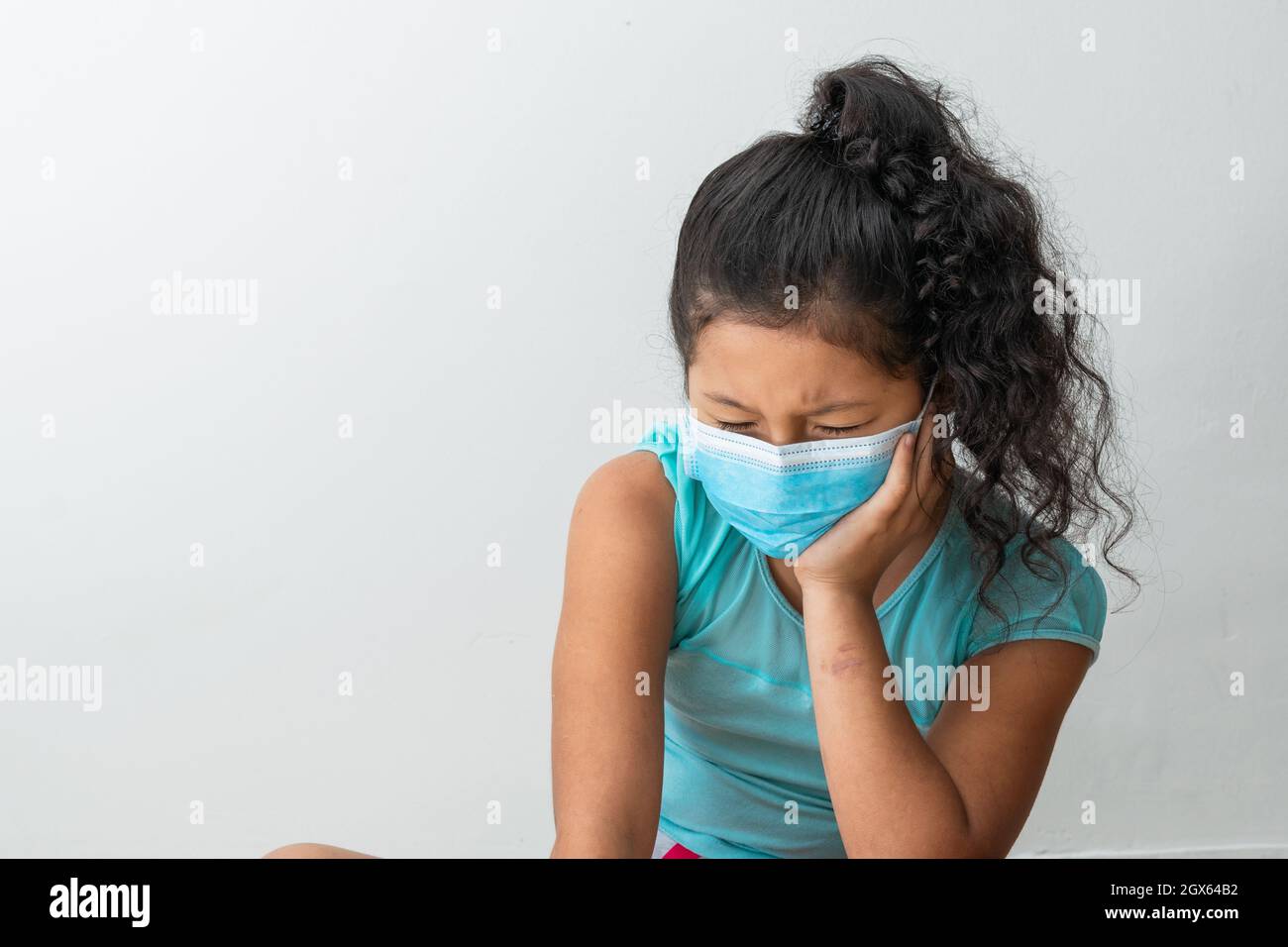 bambina (8 anni) seduta sul pavimento arrabbiata, la mano sinistra è sopra il viso e ha una maschera chirurgica blu. Medico, farmaceutico e san Foto Stock