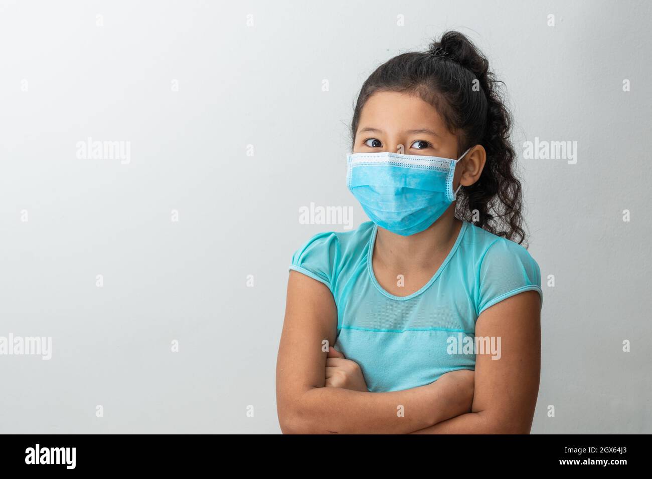 bambina (8 anni) seduta con le braccia incrociate, ragazza allegra marrone ricevendo buone notizie con una maschera chirurgica blu. Medico, farmaceutico e Foto Stock