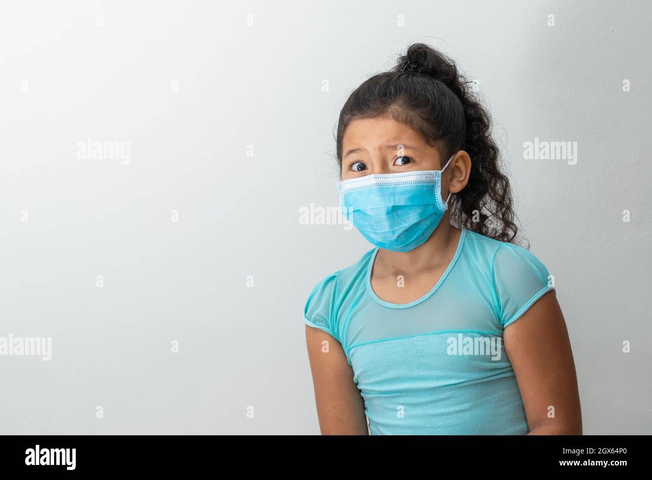 bambina (8 anni) seduta con le braccia incrociate, ragazza marrone con volto confuso che riceve una notizia con una maschera chirurgica blu. Medico, farmaceu Foto Stock