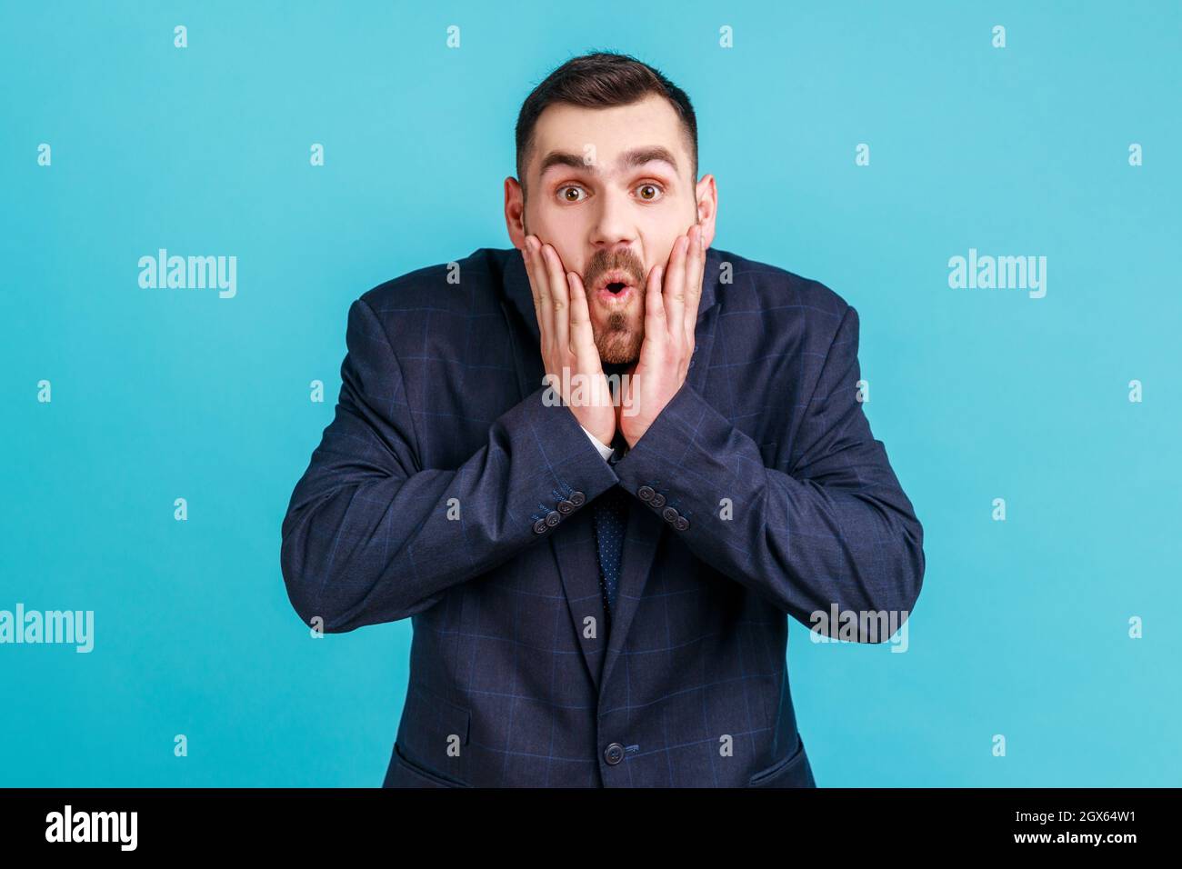 Foto di stupito giovane uomo adulto in stile ufficiale tuta guarda fotocamera a bocca aperta, alzando le palme, essendo impressionato da scioccanti notizie. Studio interno girato isolato su sfondo blu. Foto Stock