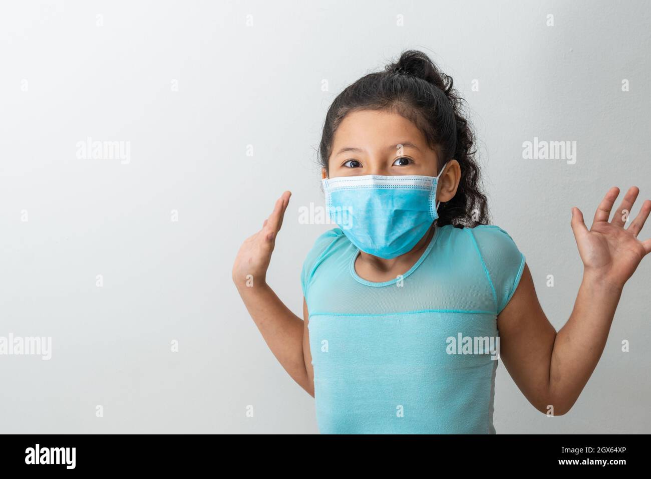 bambina (8 anni) seduta con le braccia in su, ragazza marrone che celebra la ricezione di una notizia con una maschera chirurgica blu. Medico, farmaceutico e san Foto Stock