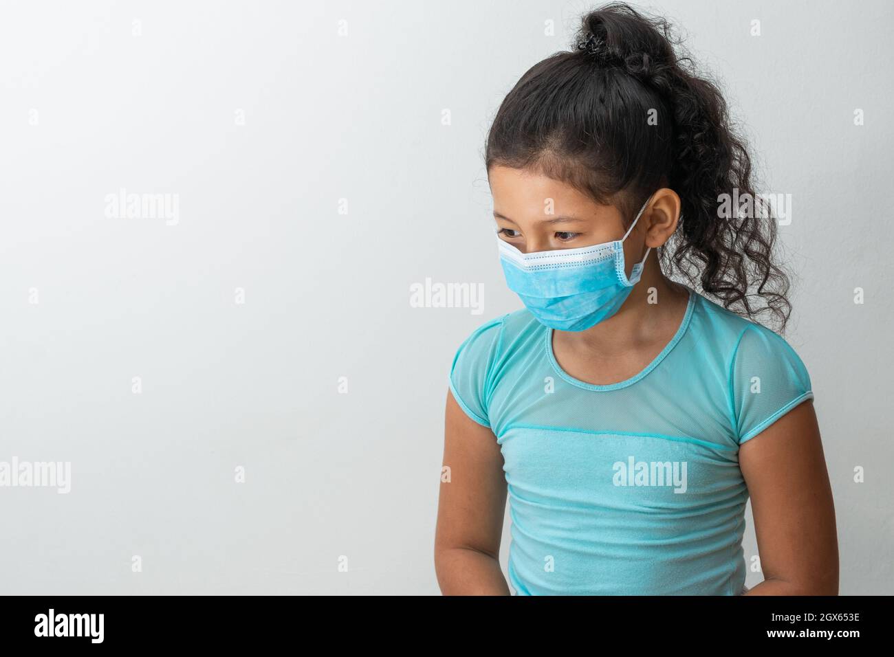 bambina (8 anni) seduta con le braccia giù confusa ragazza marrone ricevendo una notizia con una maschera chirurgica blu. Medico, farmaceutico e sanit Foto Stock