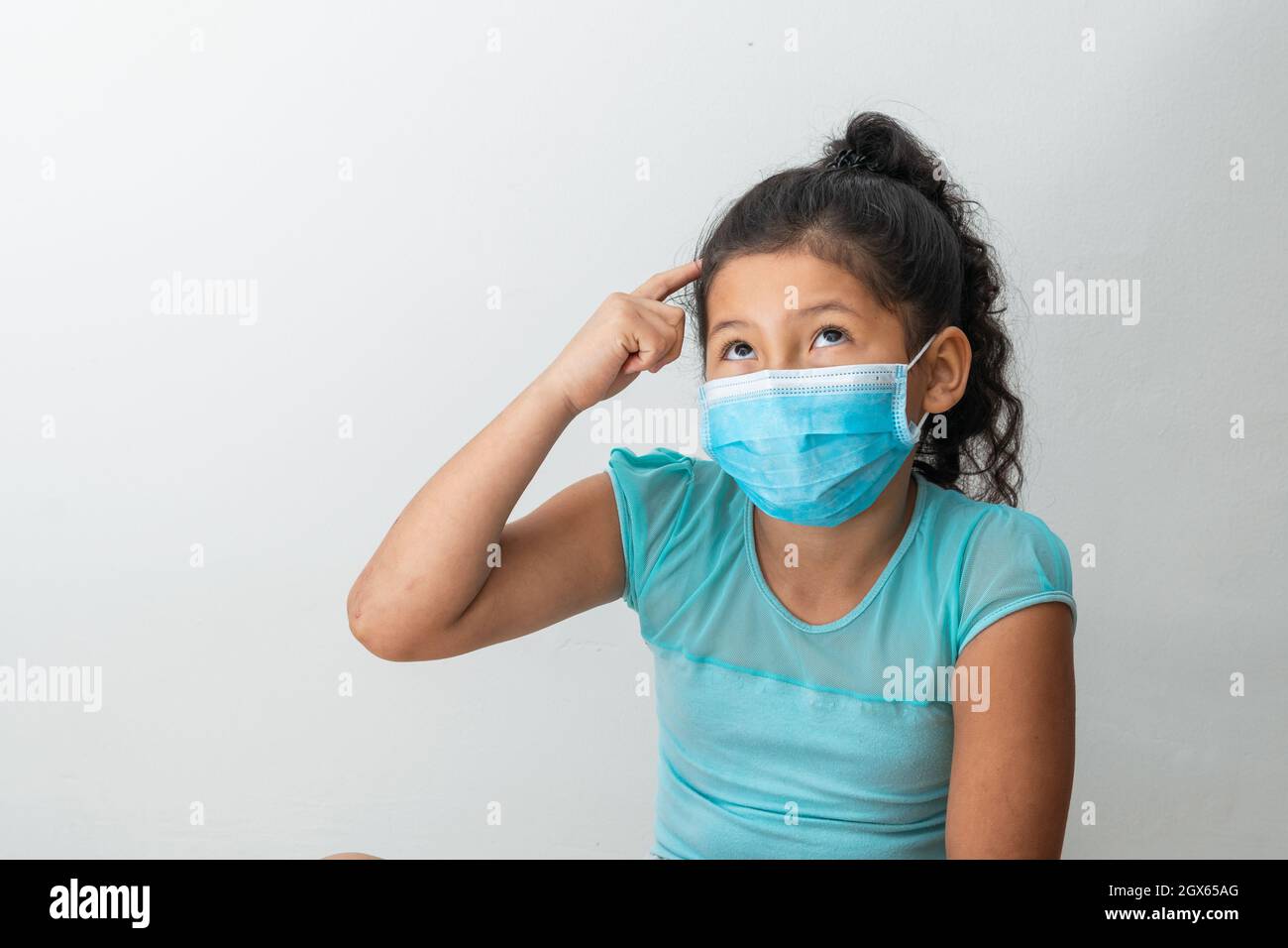 bambina (8 anni) seduta con le braccia sulla testa, ragazza marrone pensando a un'idea con una maschera chirurgica blu. Medico, farmaceutico e. Foto Stock