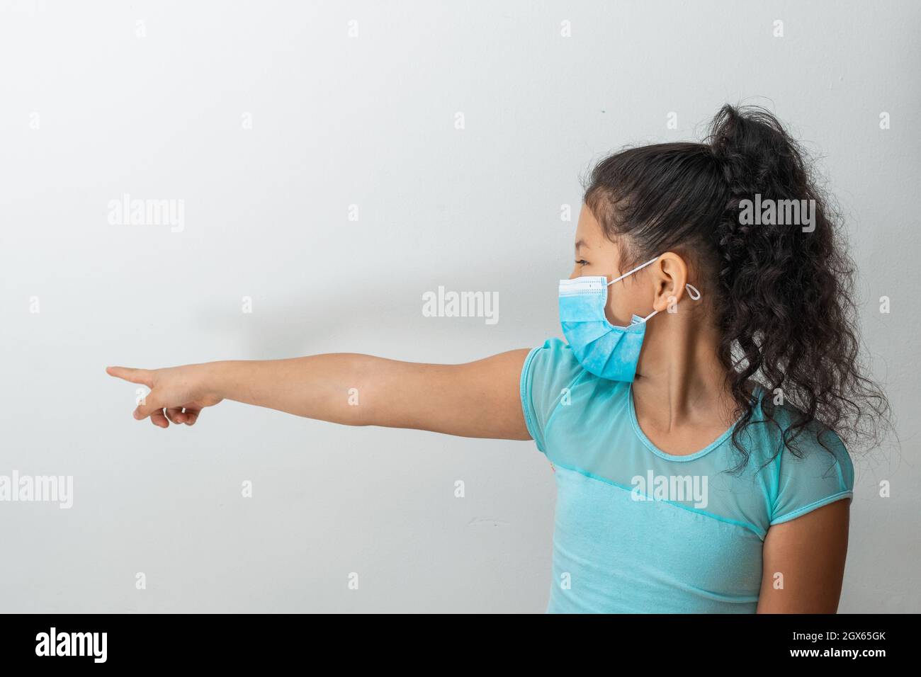 bambina (8 anni) seduta con la mano destra che punta a destra, ragazza marrone che stuzzica un'altra persona con una maschera chirurgica blu. Medico, pha Foto Stock