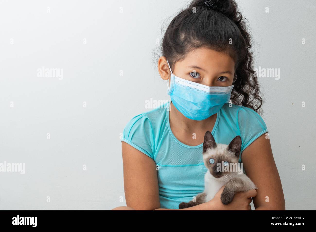 Bambina (8 anni) seduta con un gatto siamese nelle mani, ragazza marrone con una maschera chirurgica blu. Concetto medico, farmaceutico e sanitario. Foto Stock