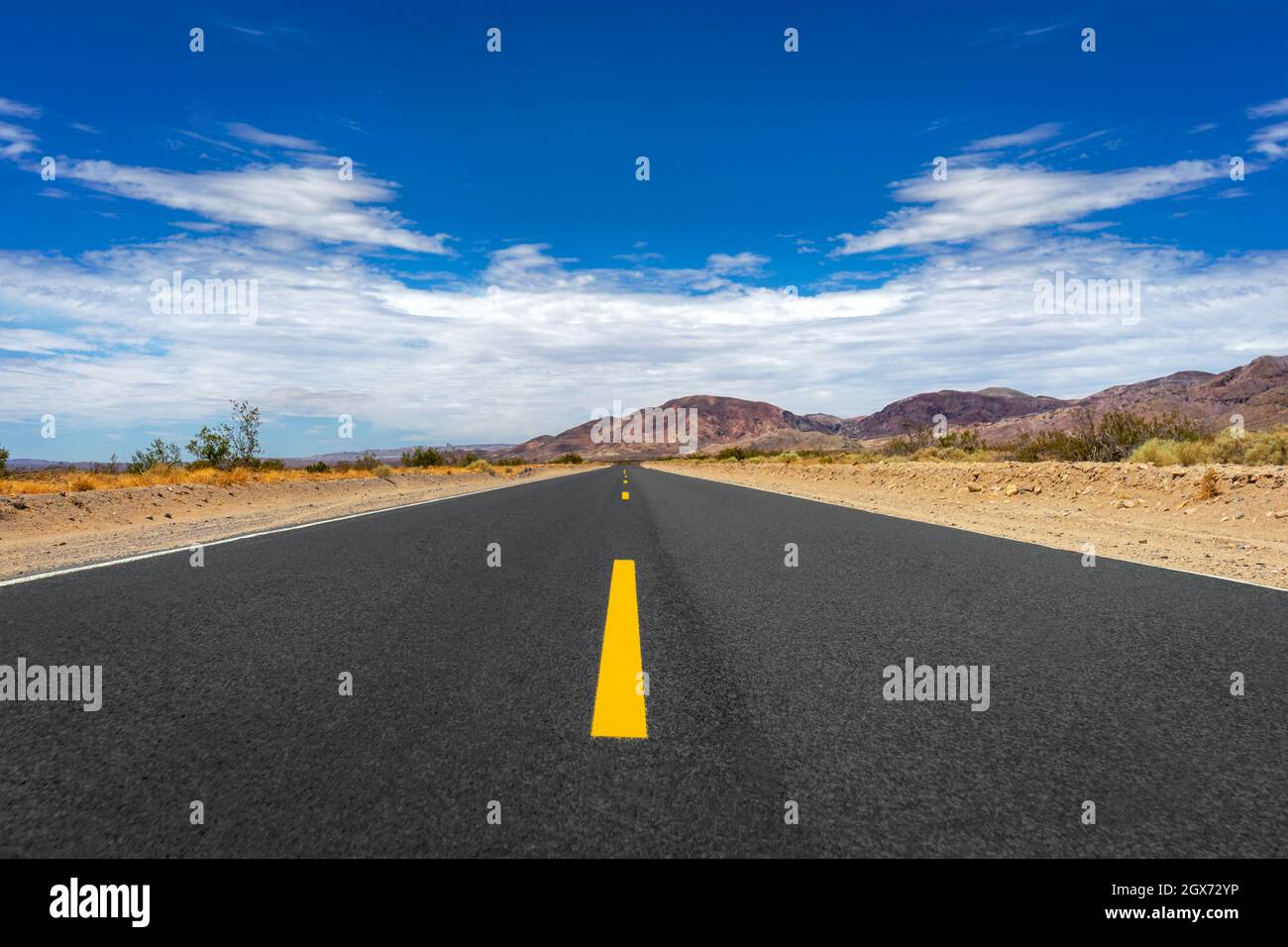 Lunga strada vuota nel deserto di Mojave in California con cielo blu nuvoloso Foto Stock