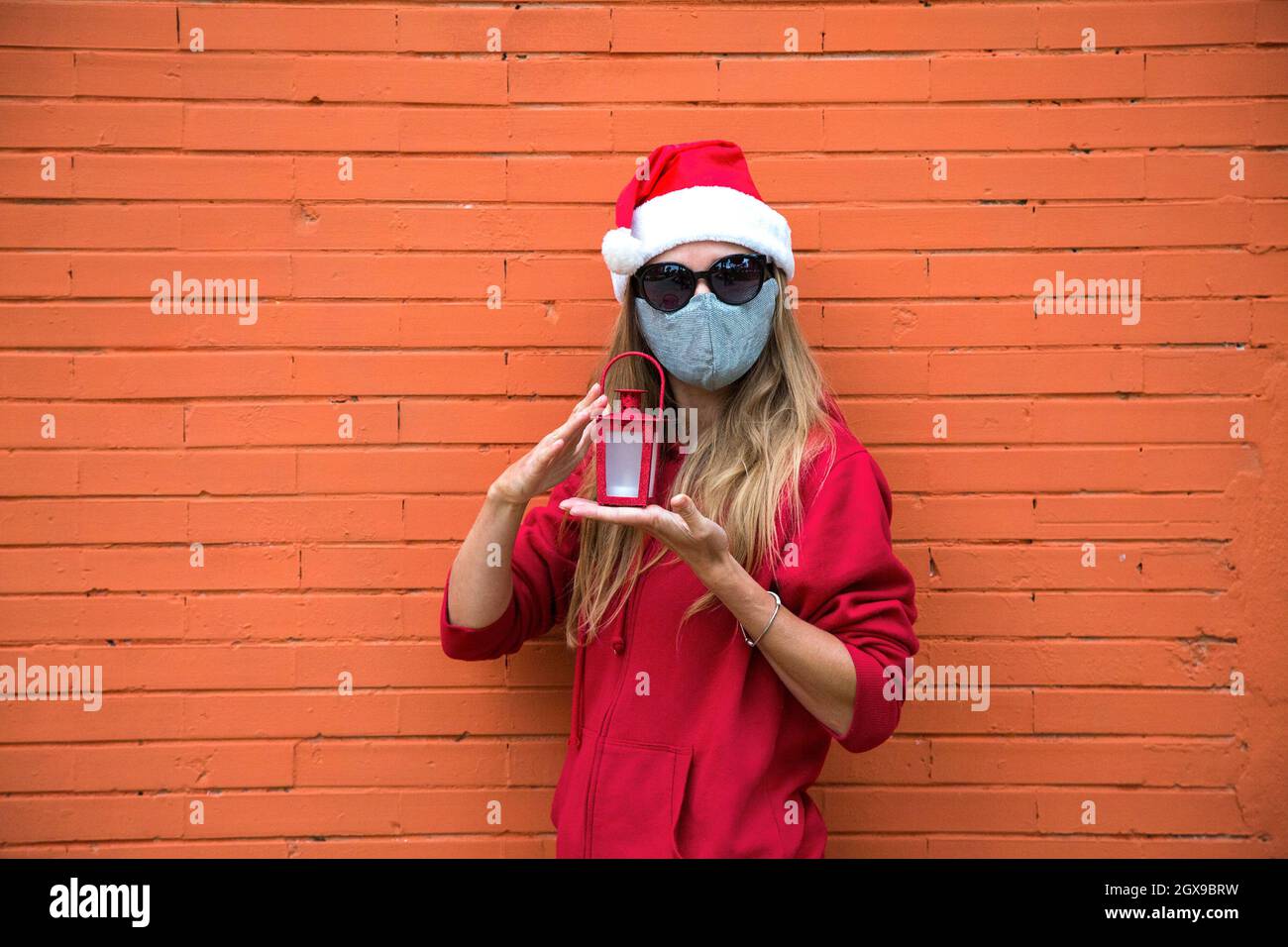 Ragazza bionda in maschera d'argento, cappello rosso e occhiali da sole neri tiene in mano la luce di Natale. Donna caucasica in costume di Natale e protezione covid Foto Stock