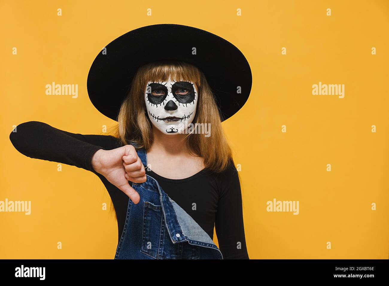 Ritratto ragazza capretto con maschera di trucco di Halloween indossa il cappello nero che dà il pollice giù il gesto che osserva con espressione negativa e disapprovazione Foto Stock