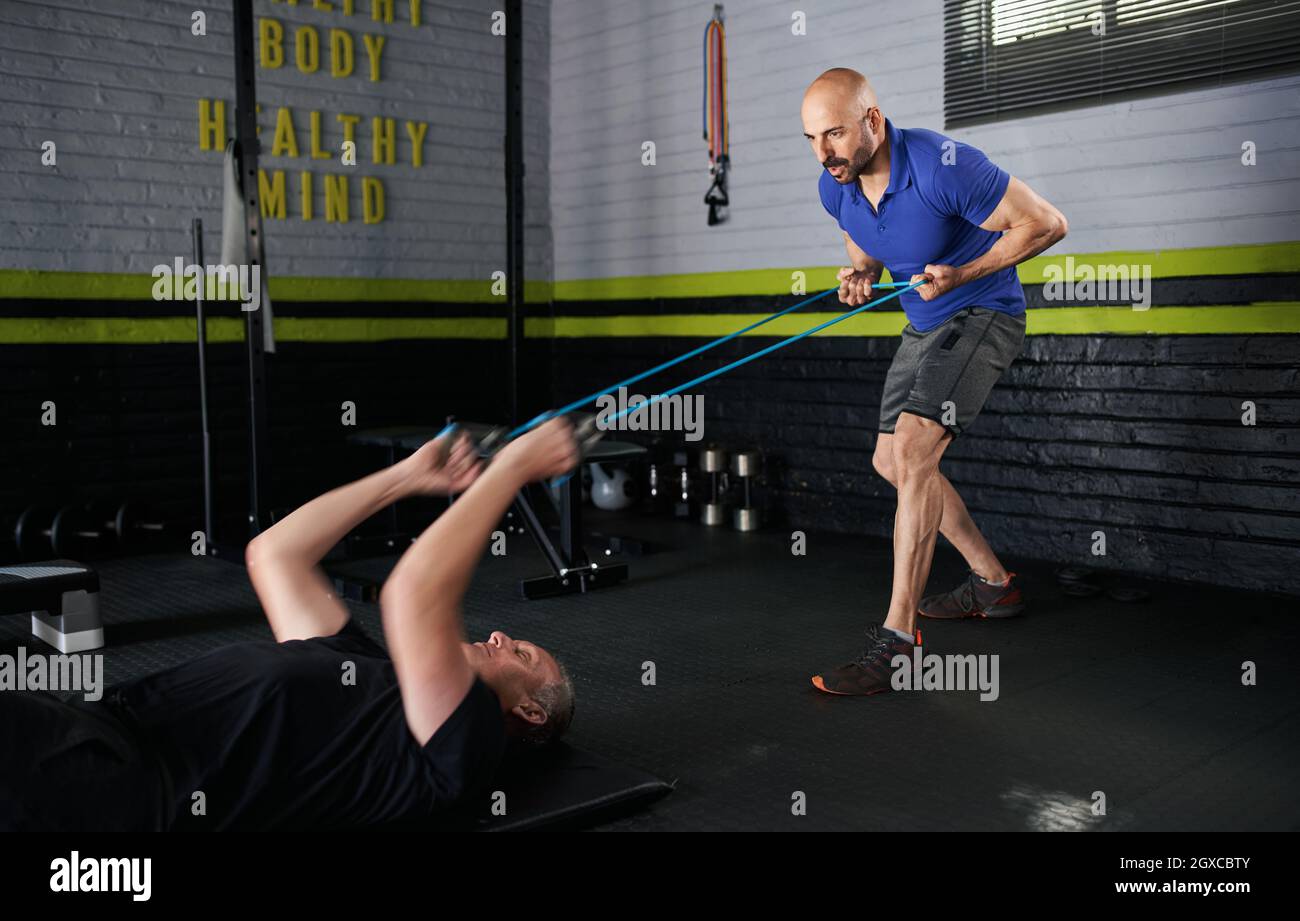 Il personal trainer maschio tiene le fasce elastiche di resistenza di gomma mentre un uomo anziano maturo sdraiato sul tappeto da sforzo sul pavimento e fa il bicep arriccia per st Foto Stock