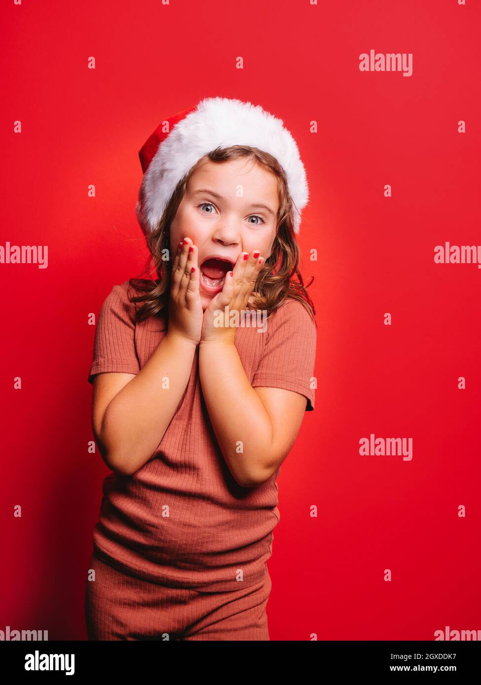 Sorprende ragazza divertente in abiti casual e cappello Santa con capelli ondulati e manicure toccare guance e urlare su sfondo rosso Foto Stock
