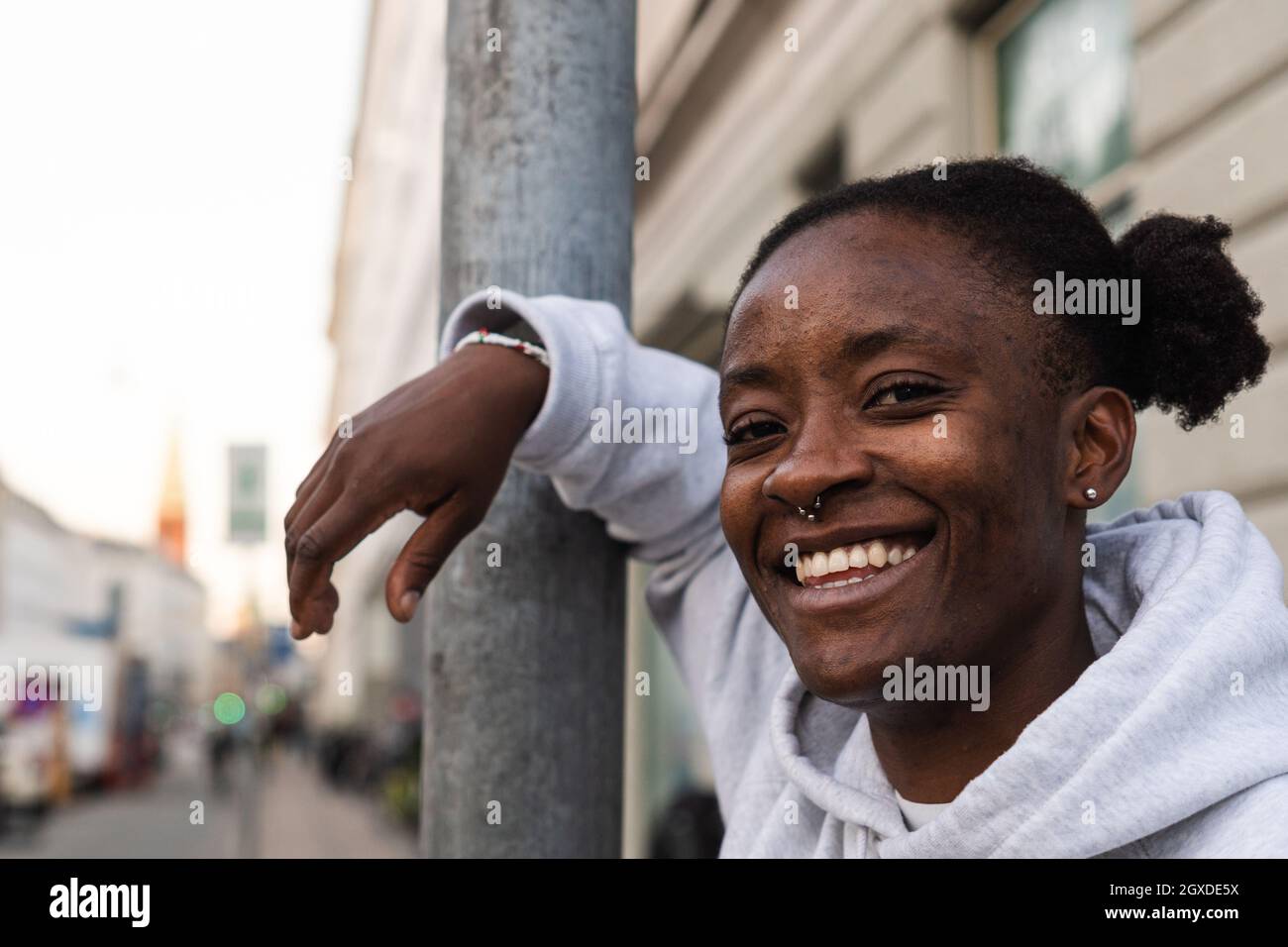 Felicissima femmina androgina afroamericana sorridente ampiamente mentre si appoggia sul pilastro di metallo e guarda la macchina fotografica Foto Stock