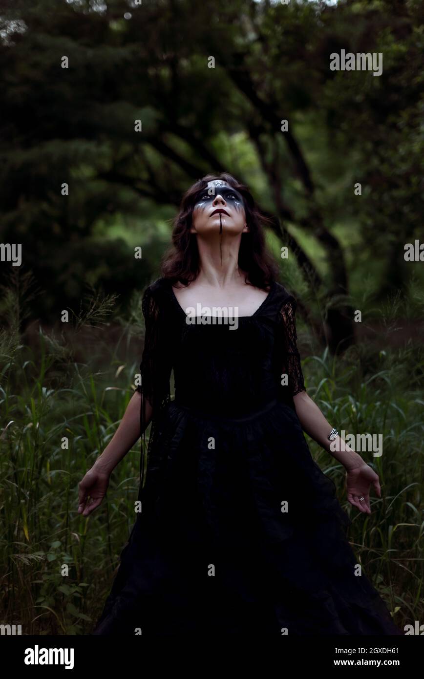 Strega mistica in lungo vestito nero e con faccia dipinta in piedi guardando in legno scuro e cupo Foto Stock