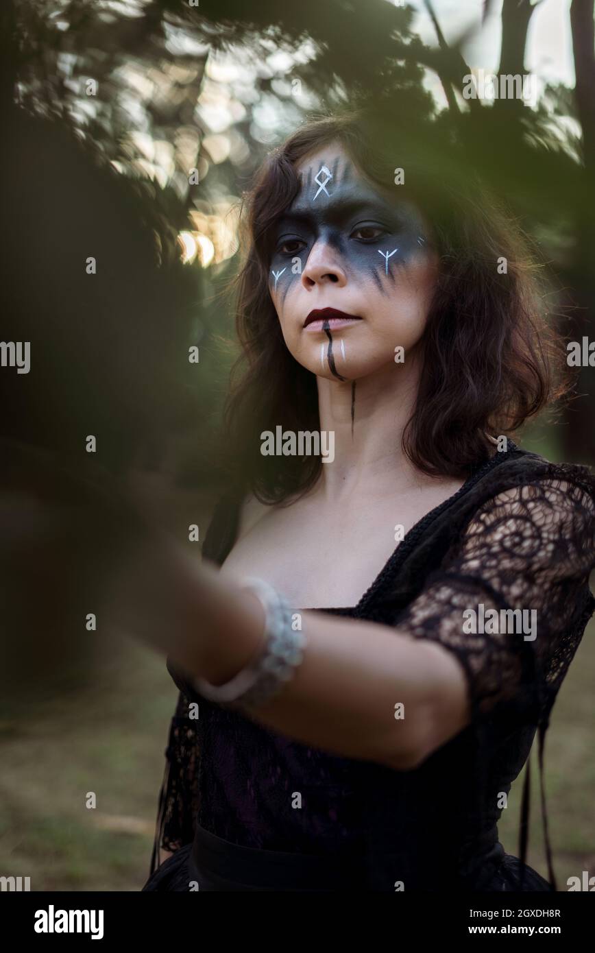Strega mistica in lungo vestito nero e con faccia dipinta in piedi e toccare foglie in legno scuro e cupo Foto Stock