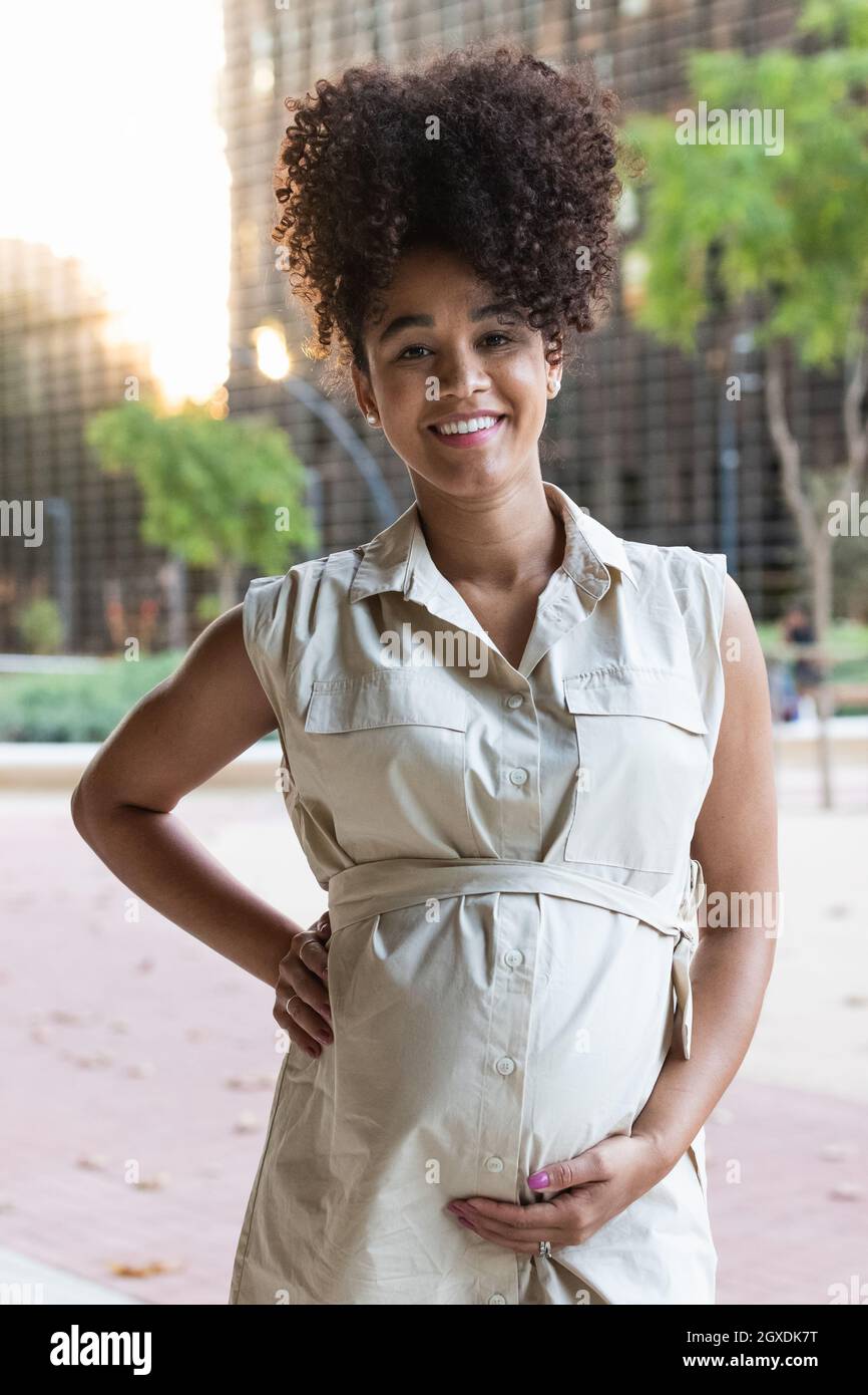 Positivo incinta femminile afroamericana in abito che tocca il ventre e guarda la macchina fotografica Foto Stock