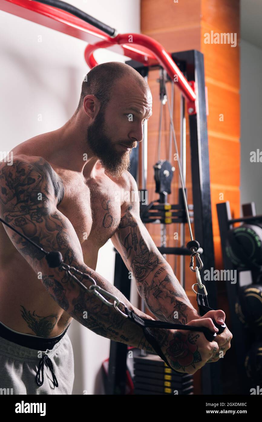 Vista laterale di sportivo muscolare concentrato con tatuaggi che fanno esercizi su cavo crossover macchina in palestra con pareti leggere Foto Stock