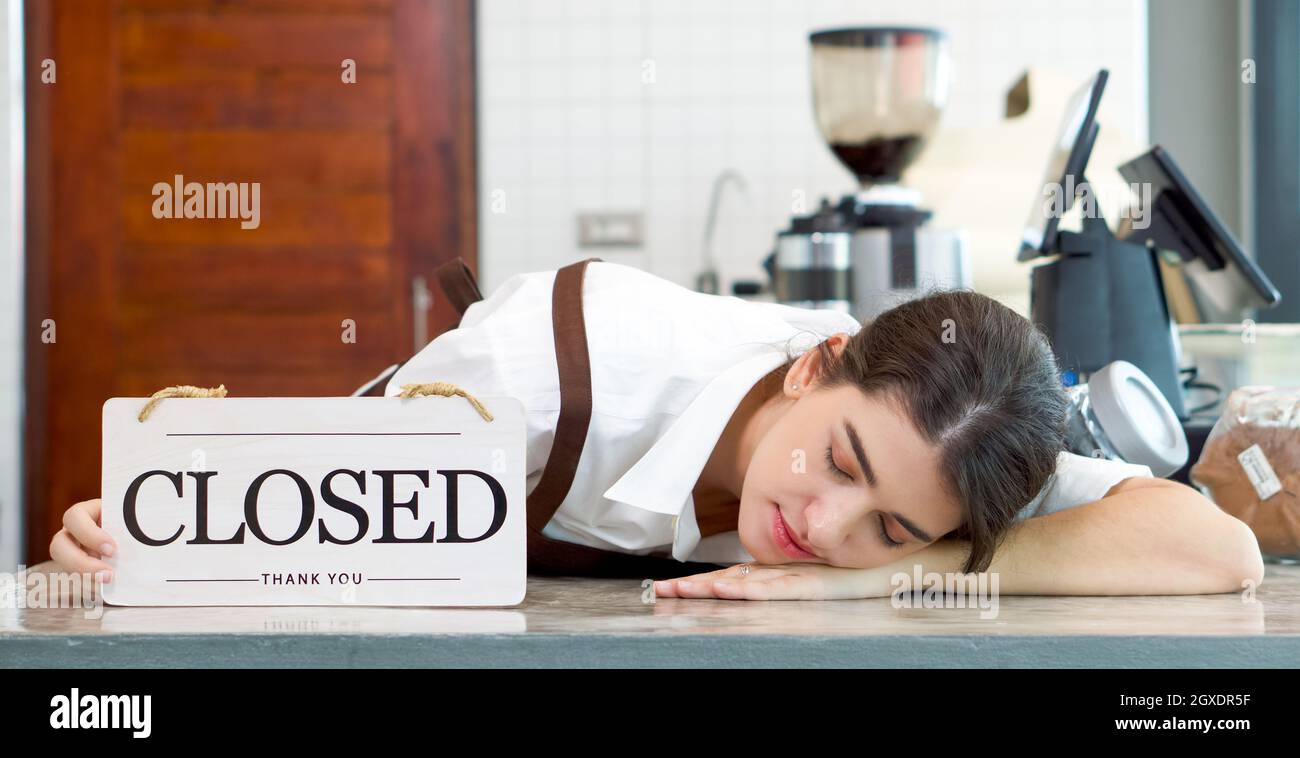 Il giovane negoziante caucasico si addormentò sul banco di una caffetteria mentre teneva un cartello di legno "chiuso". Atmosfera dopo il lavoro in una caffetteria. Foto Stock