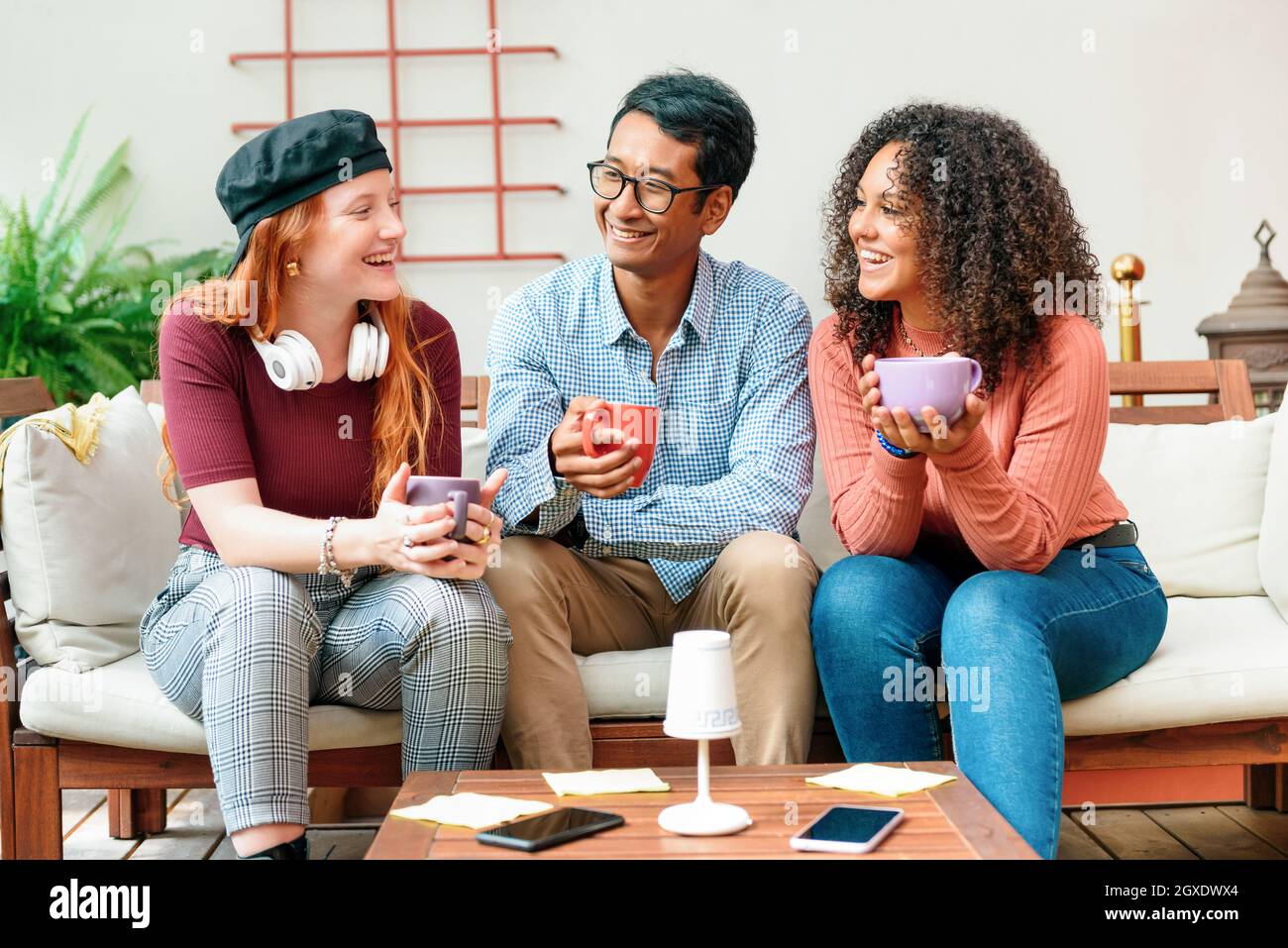Tre giovani amici diversi che si godono il caffè insieme su un divano ridendo e sorridendo, mentre trascorrono un po 'di tempo di qualità rilassarsi e chillare in un moderno Foto Stock