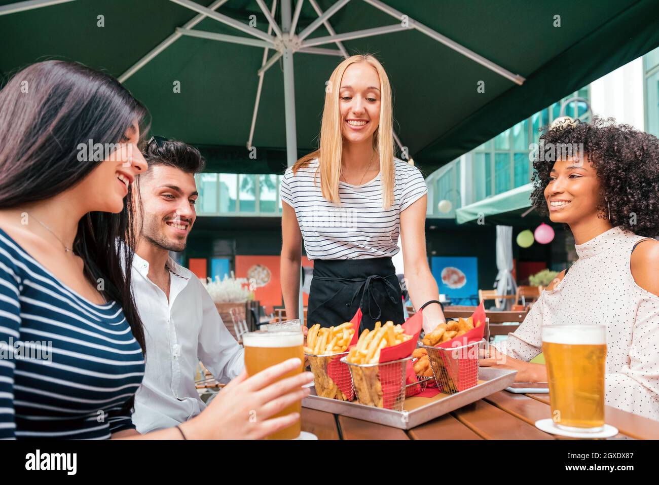 Una cameriera bionda sorridente che serve patatine fritte a un tavolo da pub a un gruppo di giovani amici diversi che si godono una birra fredda insieme Foto Stock