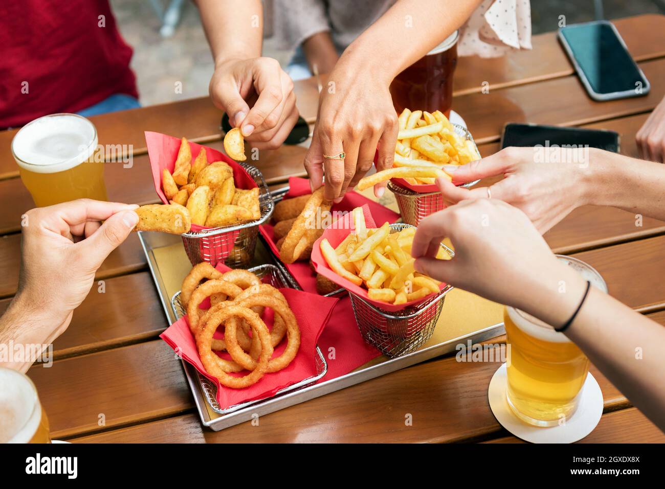 Alto angolo di gruppo di raccolto irriconoscibili amici che prendono patatine fritte e croccanti anelli di cipolla dal vassoio mentre si riuniscono e bere birra in Foto Stock