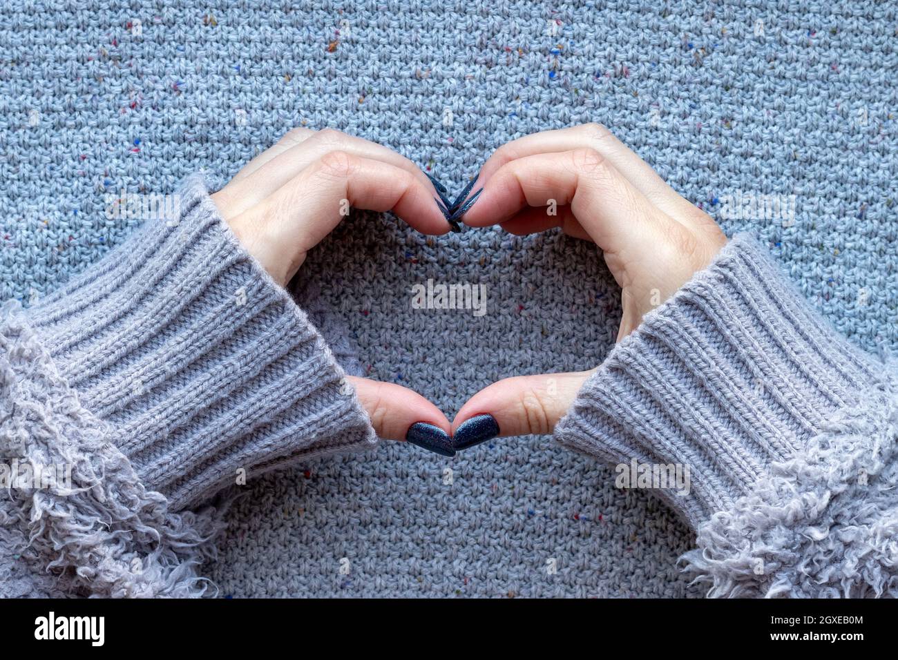 Le mani femminili in maglia grigia maglione con bella manicure - grigio scuro unghie glittered blu che fanno il simbolo del cuore su sfondo lavorato a maglia Foto Stock