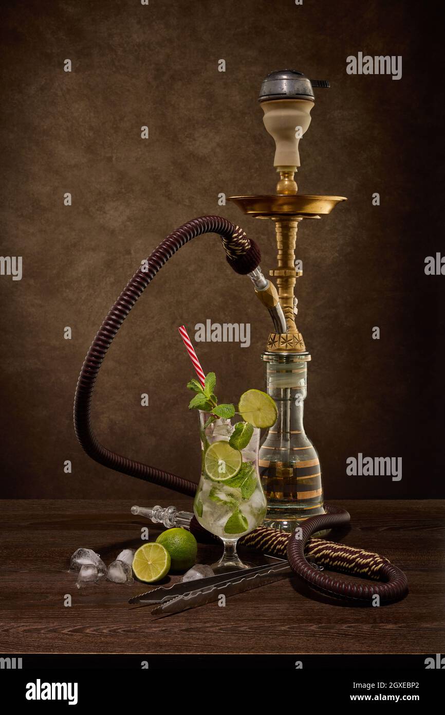 Hookah (shisha) e bicchiere di Mojito cocktail con calce e ghiaccio su tavolo di legno marrone scuro su sfondo scuro. Fine settimana o festa di vacanza Foto Stock