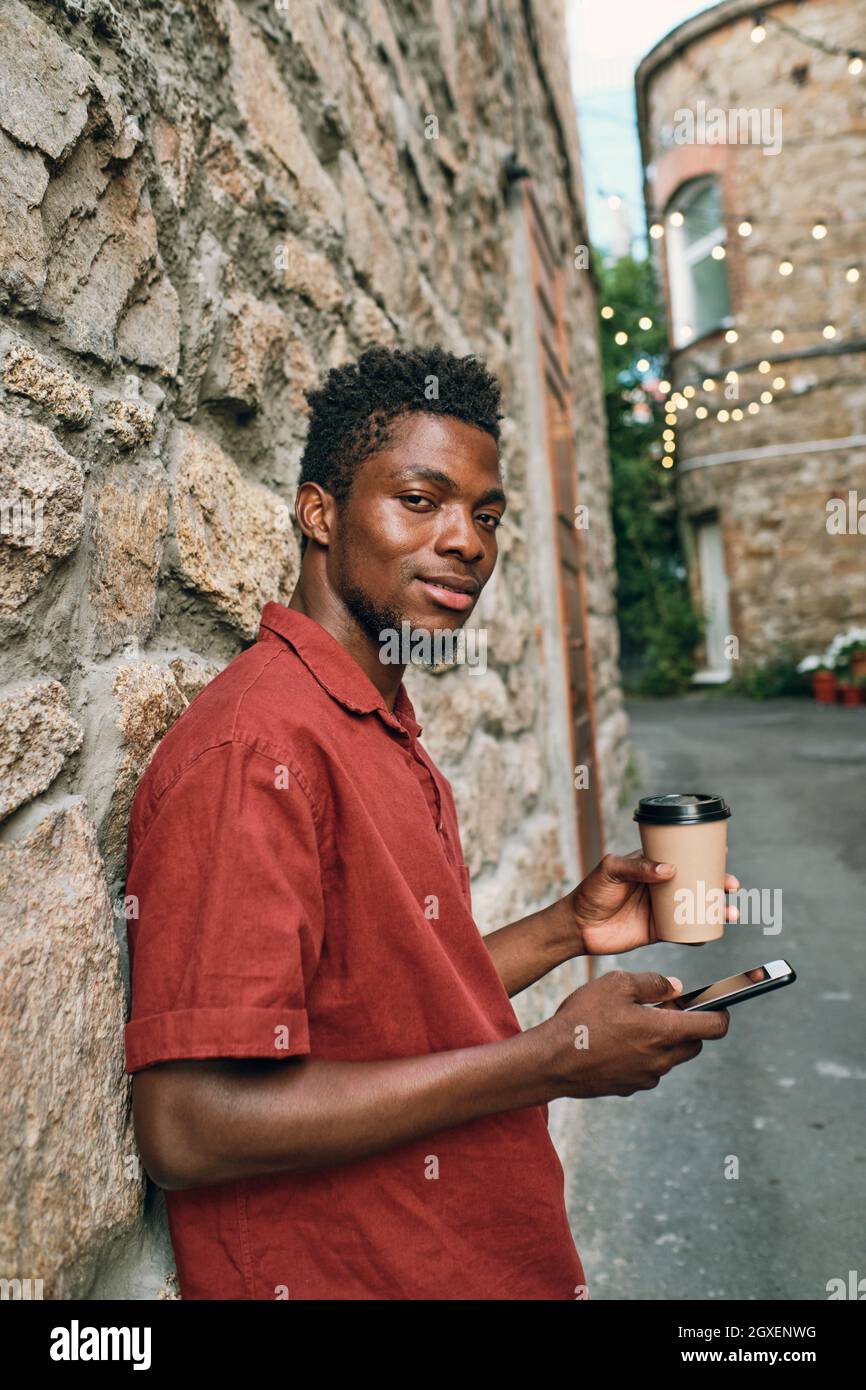 Giovane uomo di etnia africana che scorre nel cellulare e bere davanti alla macchina fotografica Foto Stock
