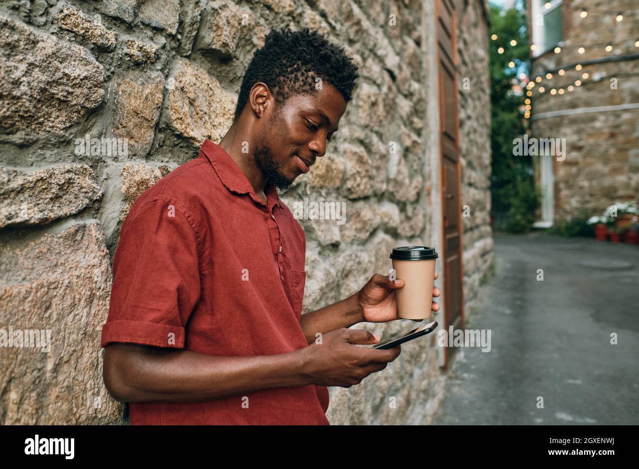 Felice giovane africano con testing bevanda in smartphone mentre si trova in piedi da muro di pietra Foto Stock