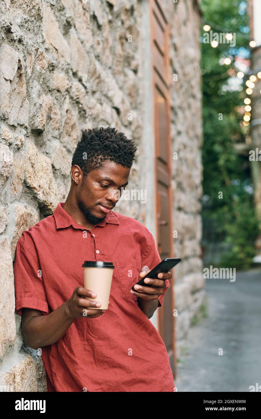 Ragazzo africano con un bicchiere di caffè guardando video online in smartphone all'aperto Foto Stock