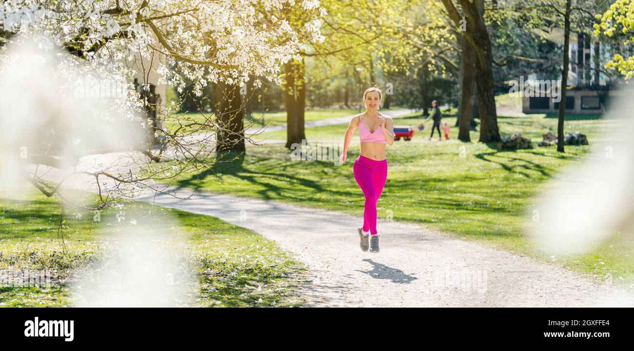 Fit donna che corre lungo un percorso verso la fotocamera durante la primavera visto attraverso la fioritura Foto Stock