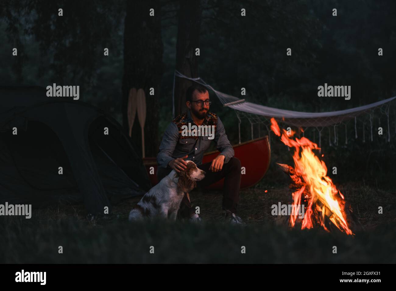 La persona si siede accanto al suo cane vicino al falò, campeggio con canoa nella natura bella Foto Stock