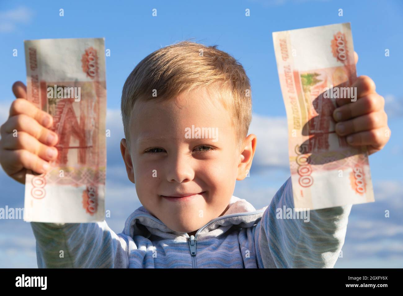 Un bambino di preschooler soddisfatto in un maglione tiene i rubli di carta nella natura in un campo contro lo sfondo di un cielo blu con le nuvole. Primo piano. Portrai Foto Stock