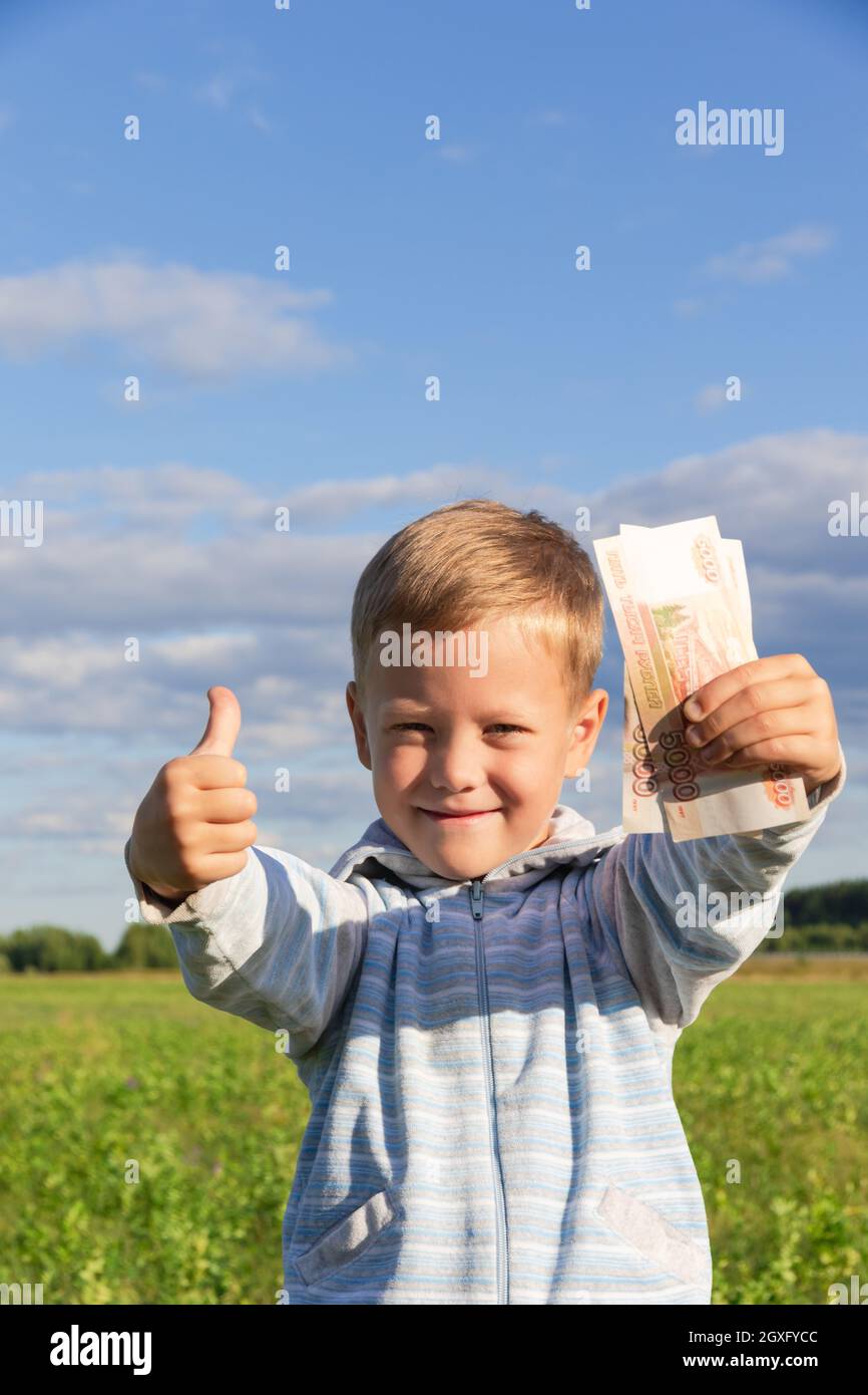 Un bambino di preschooler soddisfatto in un maglione tiene i rubli di carta nella natura in un campo contro lo sfondo di un cielo blu con le nuvole. Primo piano. Portrai Foto Stock