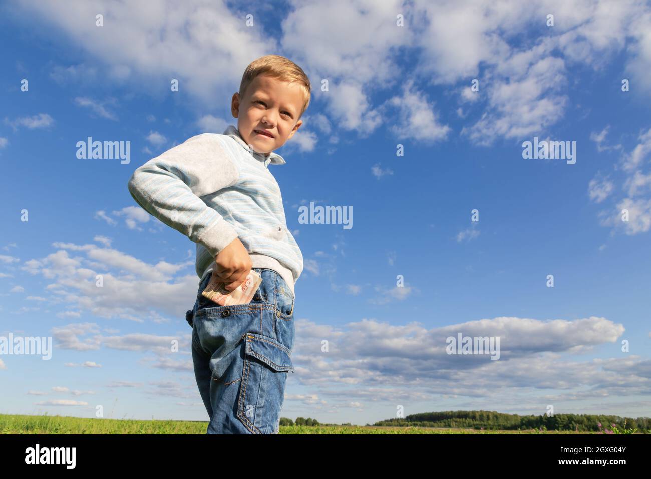 Un bambino di preschooler soddisfatto in jeans ed un maglione tiene i soldi di carta in natura in un campo contro lo sfondo di un cielo blu con le nuvole. Verticale Foto Stock