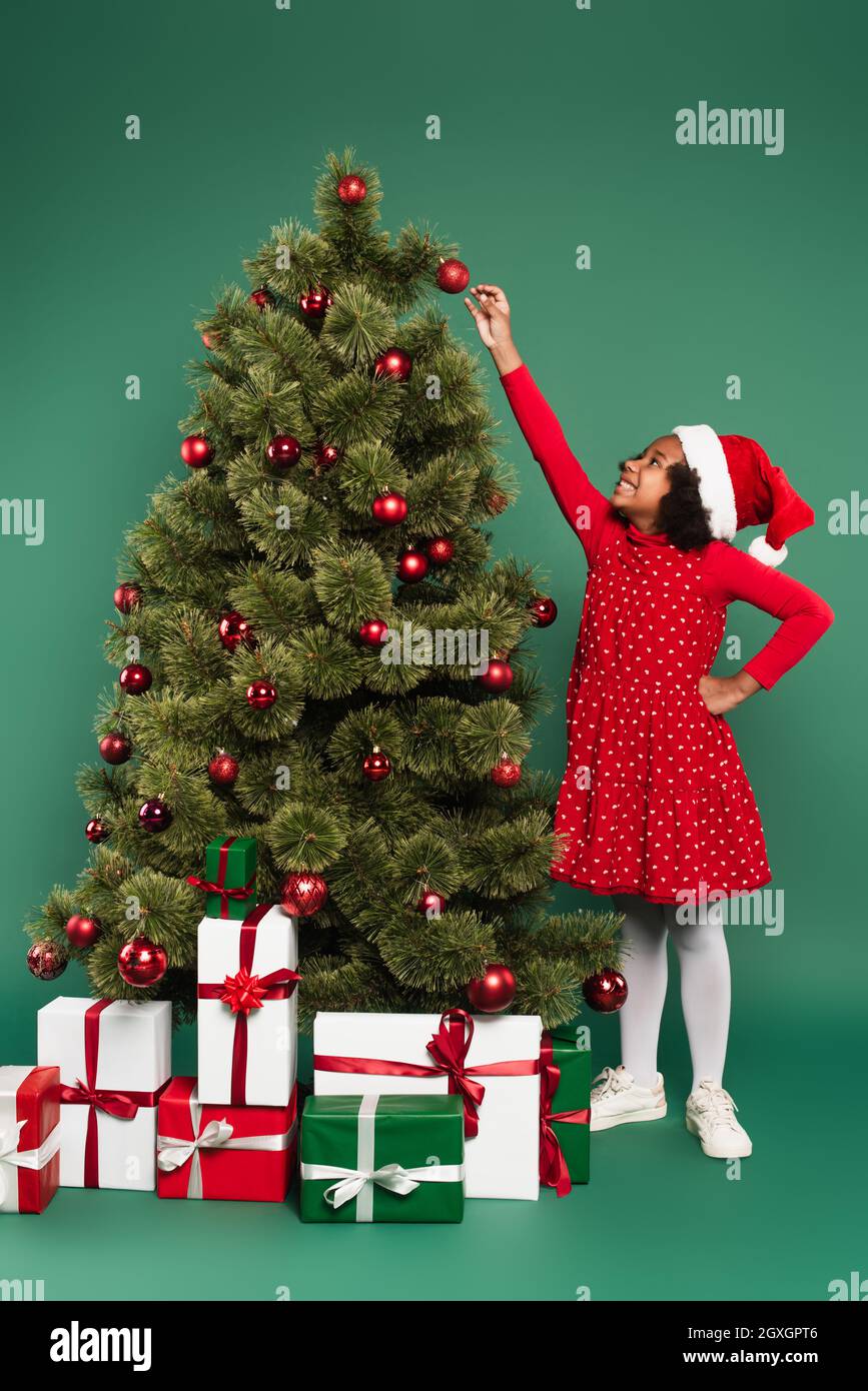 Vista laterale della ragazza afroamericana sorridente in santa Hat tirando mano a palla su albero di natale con regali su sfondo verde Foto Stock
