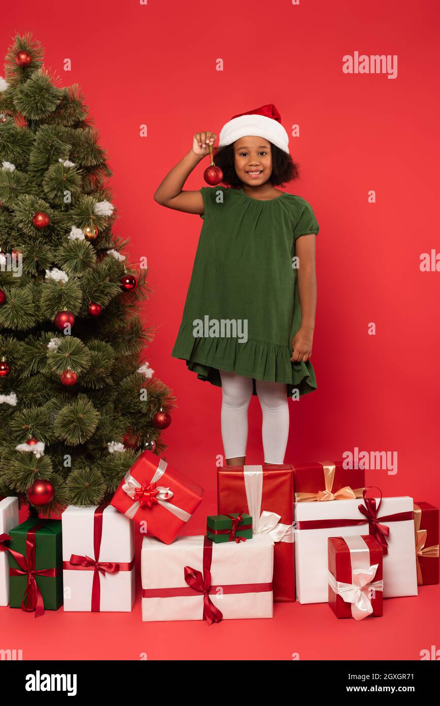 Sorridente bambino afroamericano a santa Hat che tiene la palla di natale vicino a regali e abete albero su sfondo rosso Foto Stock