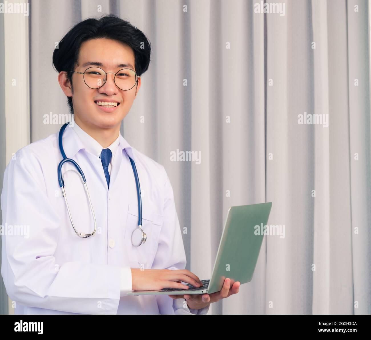 Ritratto del giovane bel dottore asiatico che indossa un vestito medico e stetoscopio sorridente seduto in ospedale, concetto di assistenza sanitaria Foto Stock