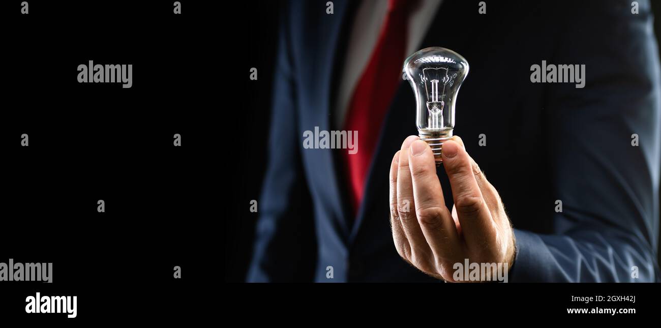 Uomo d'affari in abito nero tiene una lampadina in mano su uno sfondo nero con spazio copia. Foto Stock