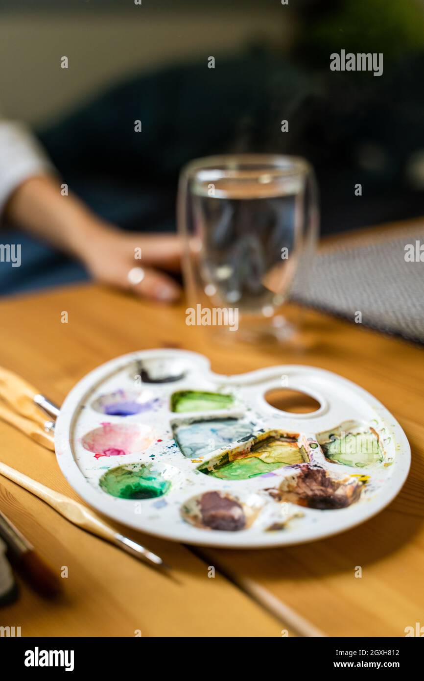 Ragazza vicino acquerelli, pennelli e tavolozze sul tavolo accanto a acqua calda in vetro Foto Stock