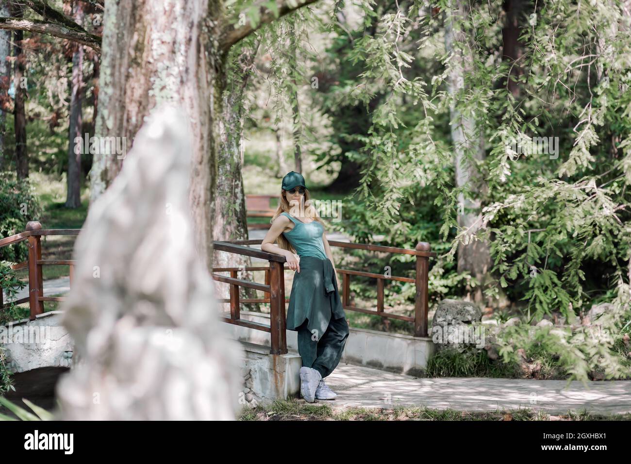 Sorridente ragazza sportiva in abito smeraldo si erge con il gomito poggiato sulla ringhiera del ponte nel verde parco estivo Foto Stock
