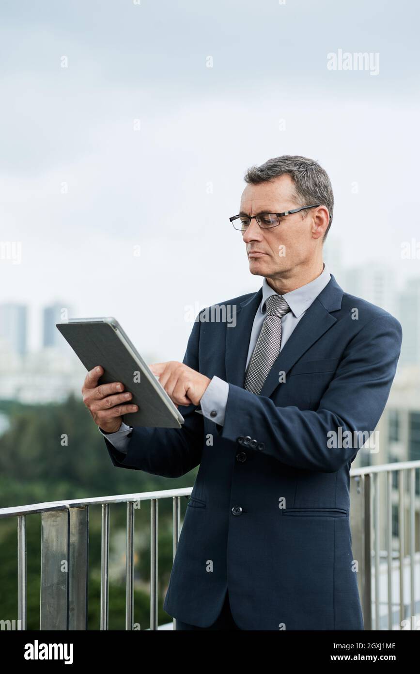 Concentrato uomo d'affari caucasico maturo in abito scuro in piedi sul balcone e tablet digitale usin Foto Stock