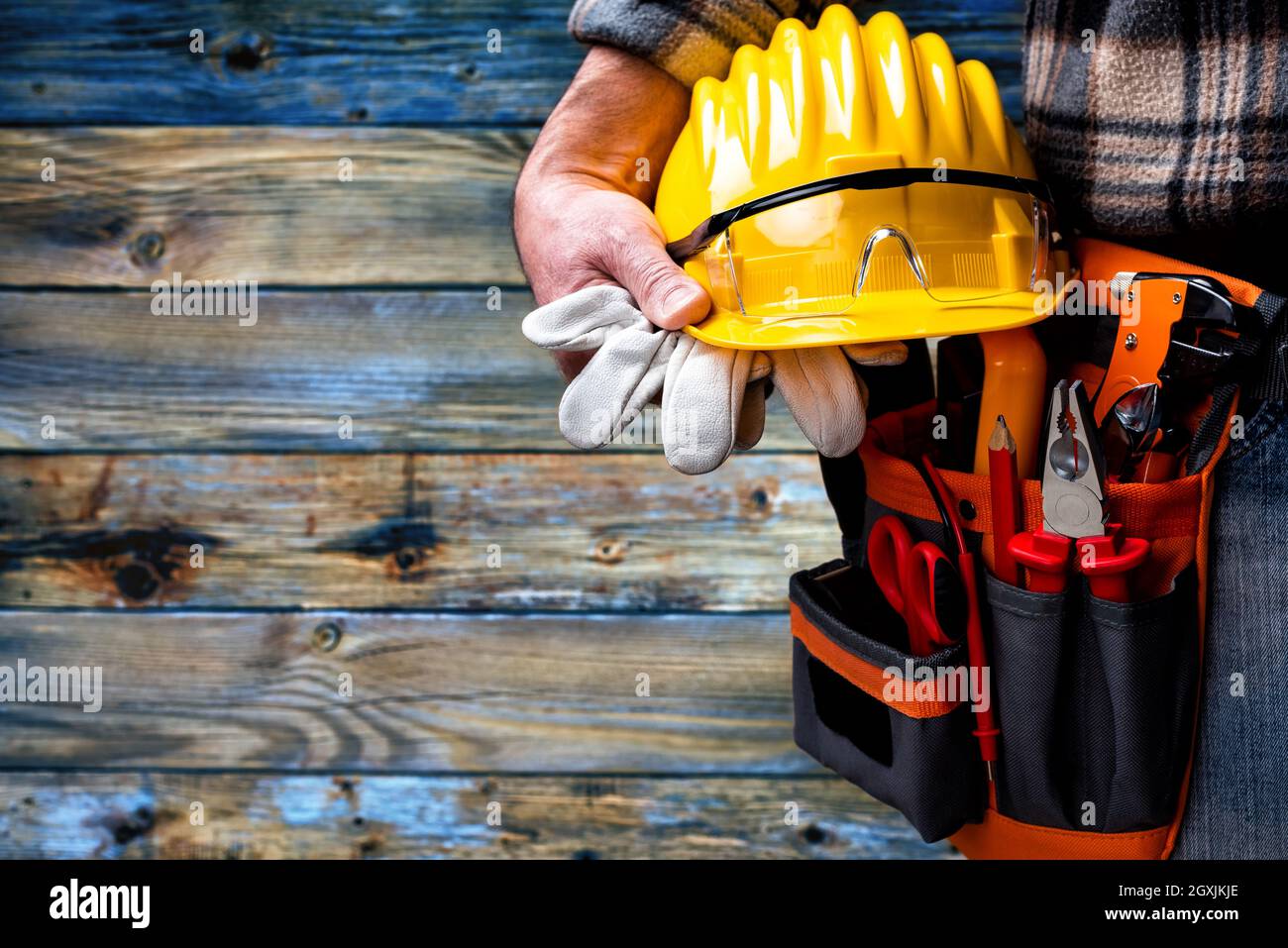 Elettricista su sfondo di legno blu chiaro vintage; tiene casco, guanti e occhiali di protezione in mano. Industria edile, impianto elettrico. Foto Stock