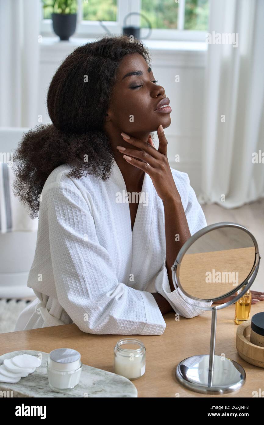 Giovane ragazza di bellezza nera che tocca il viso di fronte allo specchio in bagno a casa. Foto Stock