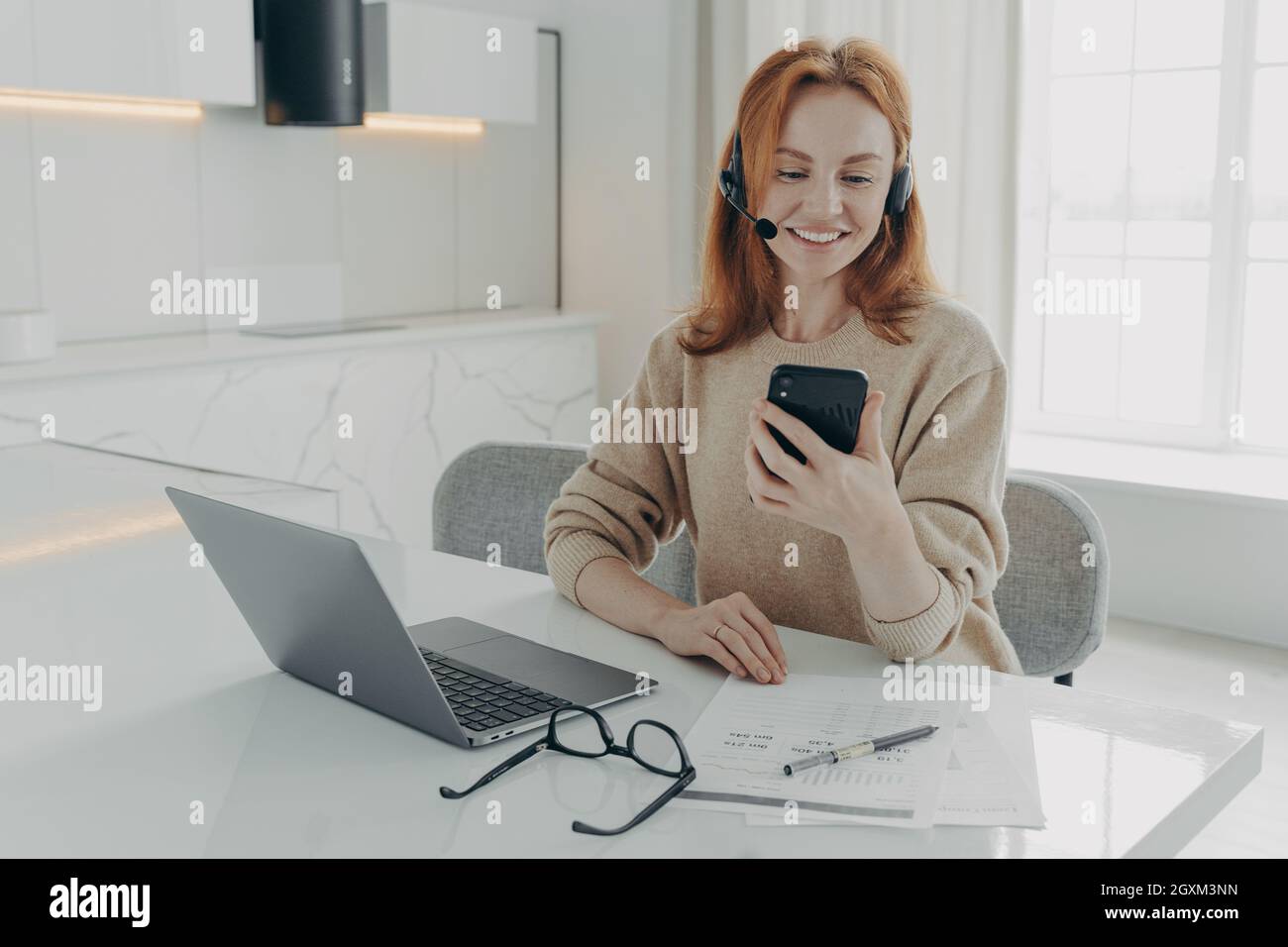 Sorridendo una donna che lavora in ufficio in cuffia utilizzando lo smartphone mentre si siede al suo posto di lavoro con un computer portatile Foto Stock
