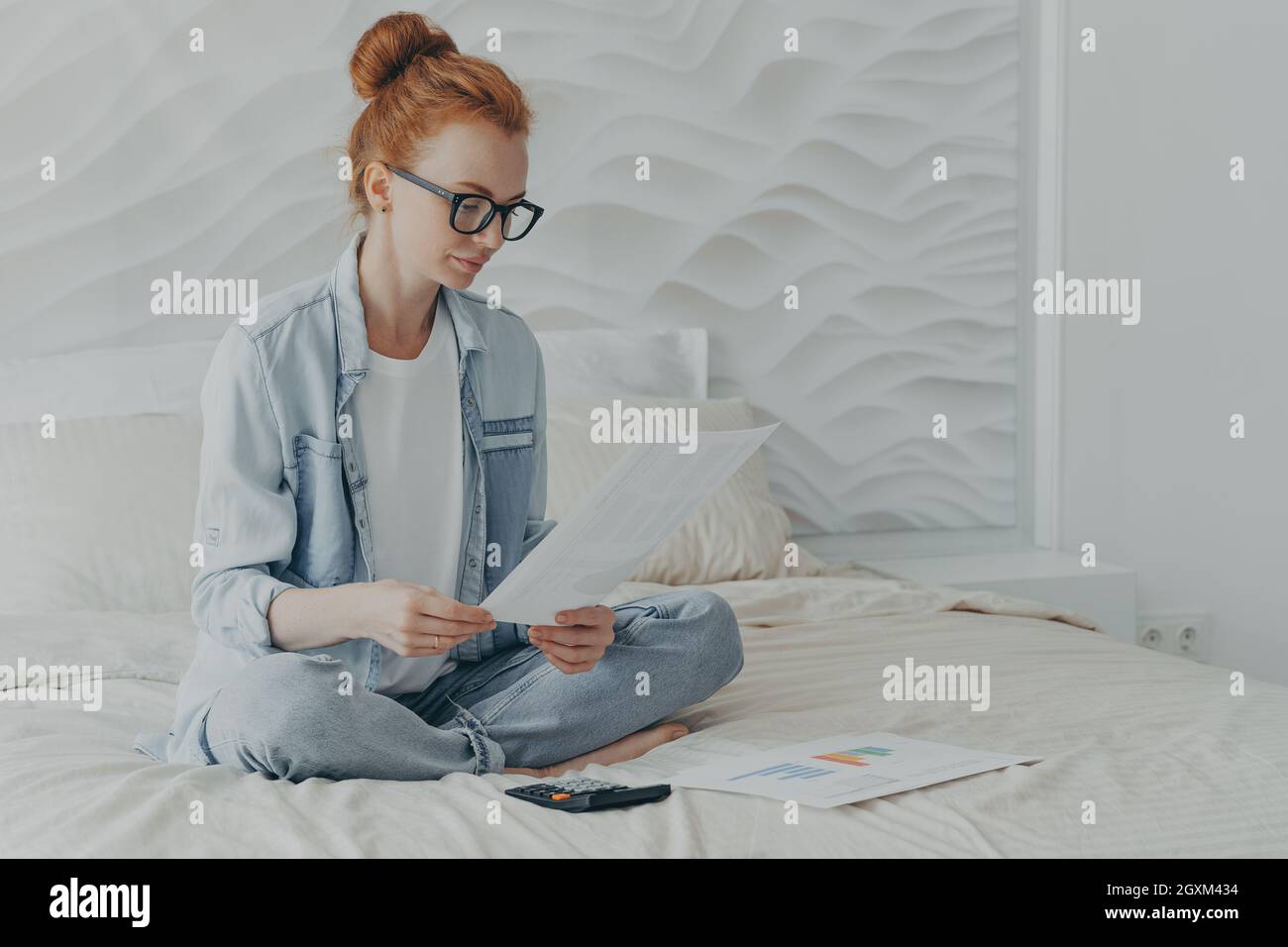 La giovane donna siede gambe incrociate su comodo letto controlla i documenti finanziari calcola il budget della famiglia Foto Stock