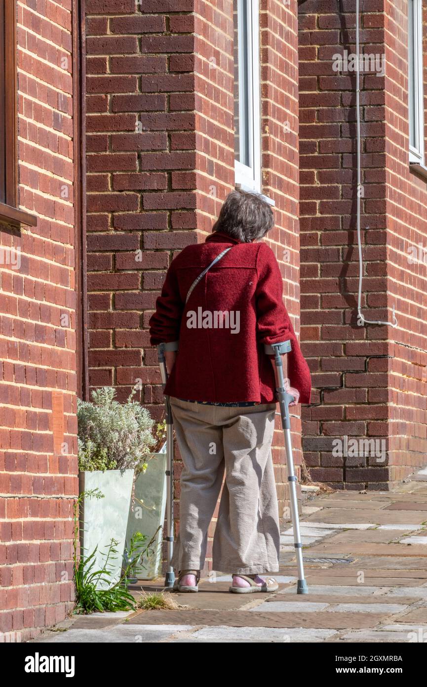 signora anziana sulle stampelle che lottano per camminare su una collina vicino ad alcune case di mattoni rossi. signora anziana disabile con stampelle a piedi sulla collina. Foto Stock