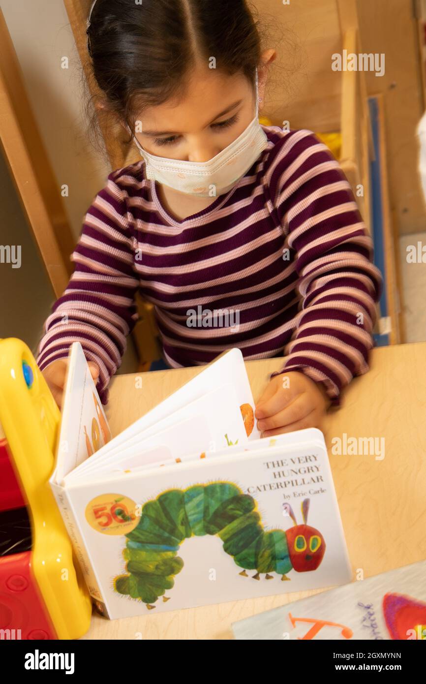 Istruzione Preschool 3-4 year olds girl looking at board book Eric Carle's The very Hungry Caterpillar wearing face mask Foto Stock
