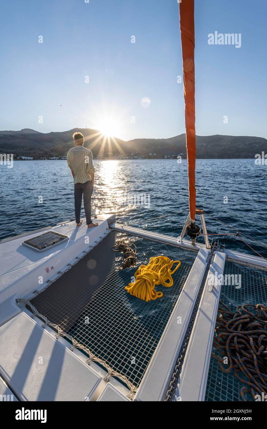 Giovane uomo in piedi su un catamarano che guarda al tramonto, corde su un catamarano a vela, stella del sole, gita in barca a vela, Tilos, Dodecaneso, Grecia Foto Stock
